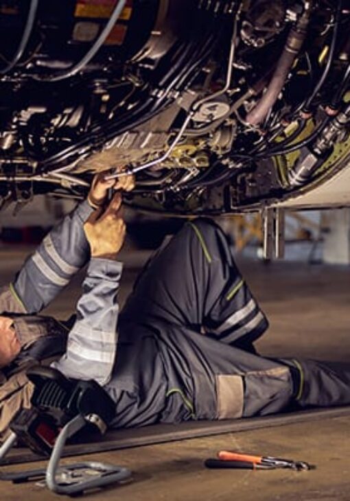 Aircraft maintenance technician lying on the ground under a plane, using tools to repair the underside of the engine in a hangar.