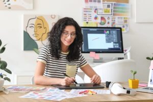 Smiling woman using a pen tablet at a desk with color swatches, working on digital art or design in a bright, creative workspace.