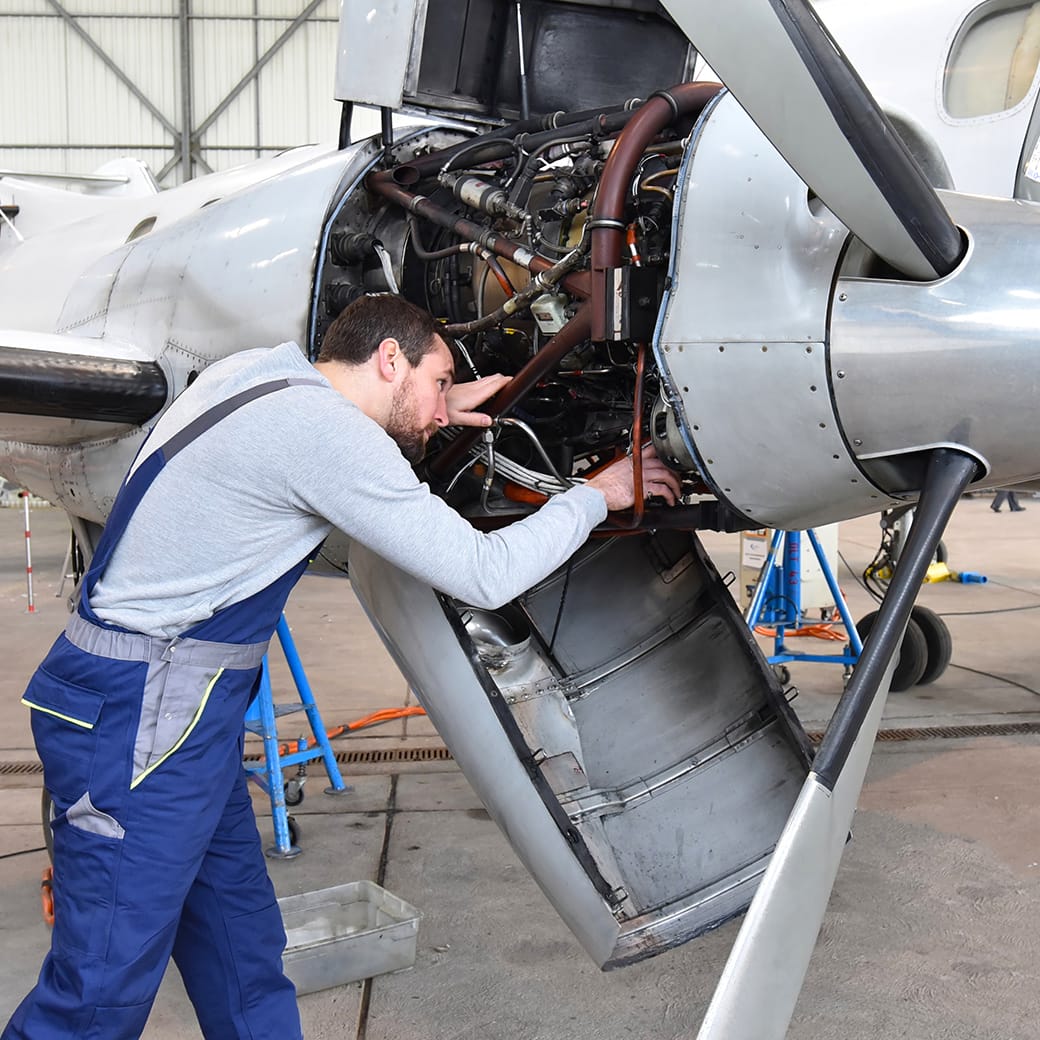 Male technician inspecting and repairing the engine of a propeller aircraft inside a hangar.