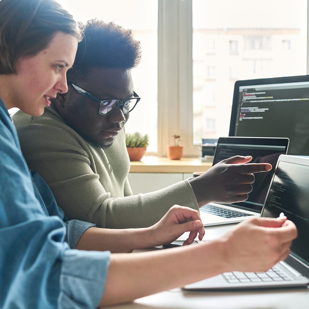 Two people collaborating on code at a desk with laptops, one pointing at the screen while the other types.