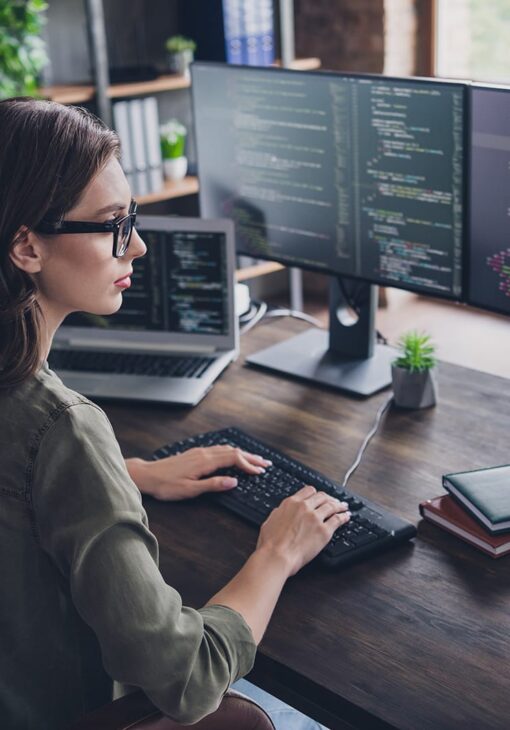 Woman with glasses coding at a desk with multiple monitors and a laptop in a modern office space.