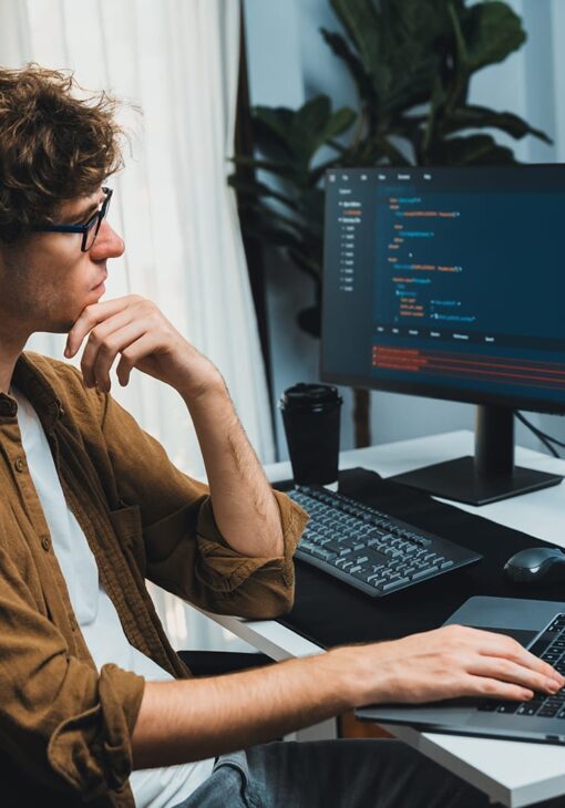 Man in glasses thoughtfully looking at code on a monitor while working on a laptop at a desk.