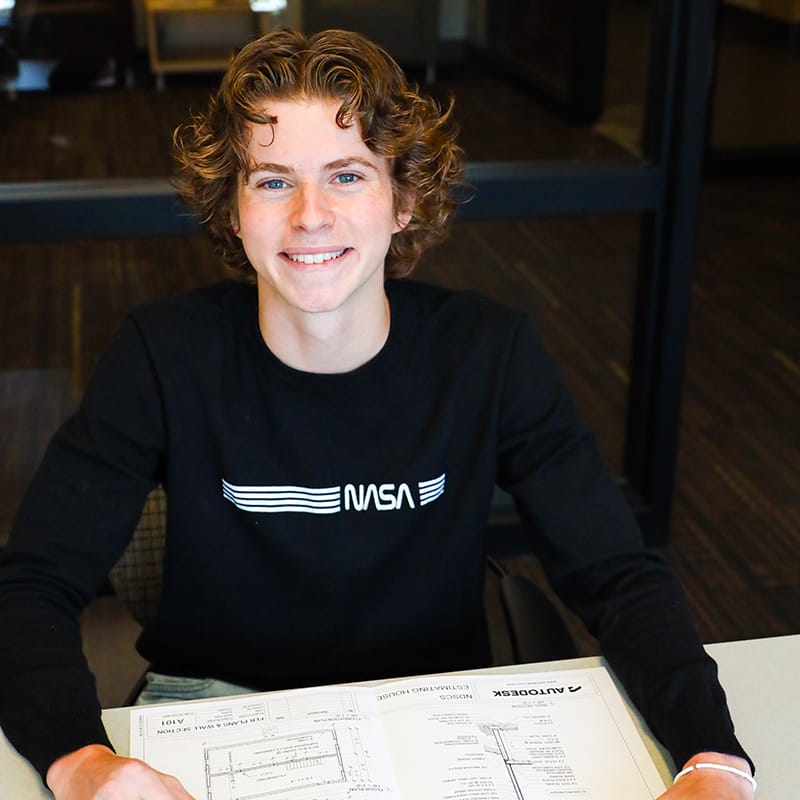 A person smiling, wearing a NASA shirt, sitting at a table with an open blueprint.