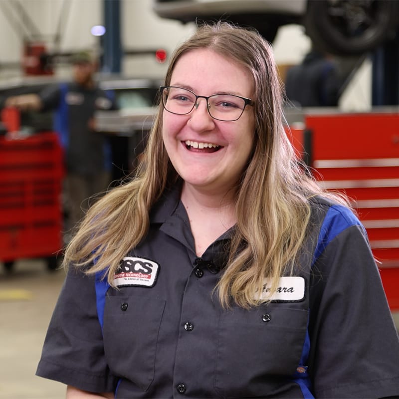 Smiling young woman in an automotive technology uniform with name tag “Amara,” standing in a brightly lit auto shop with red tool chests and vehicles in the background.