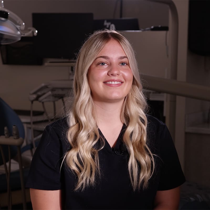 Smiling woman with long blonde hair in black scrubs sits in a dental clinic exam room.