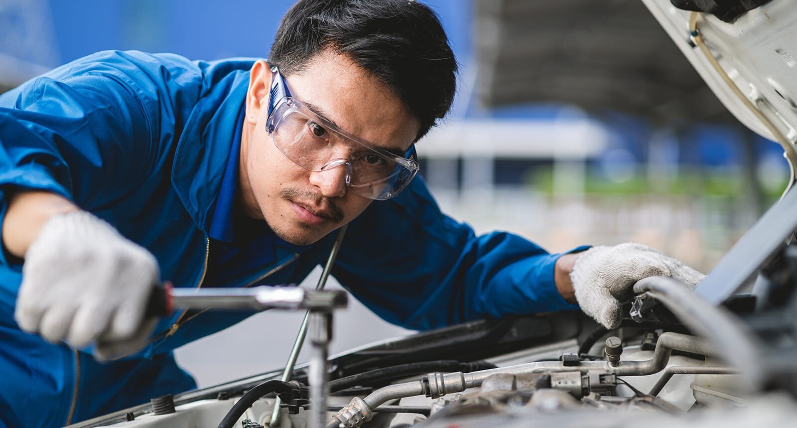 Focused automotive technician in blue coveralls and safety goggles using a wrench to repair a vehicle engine.