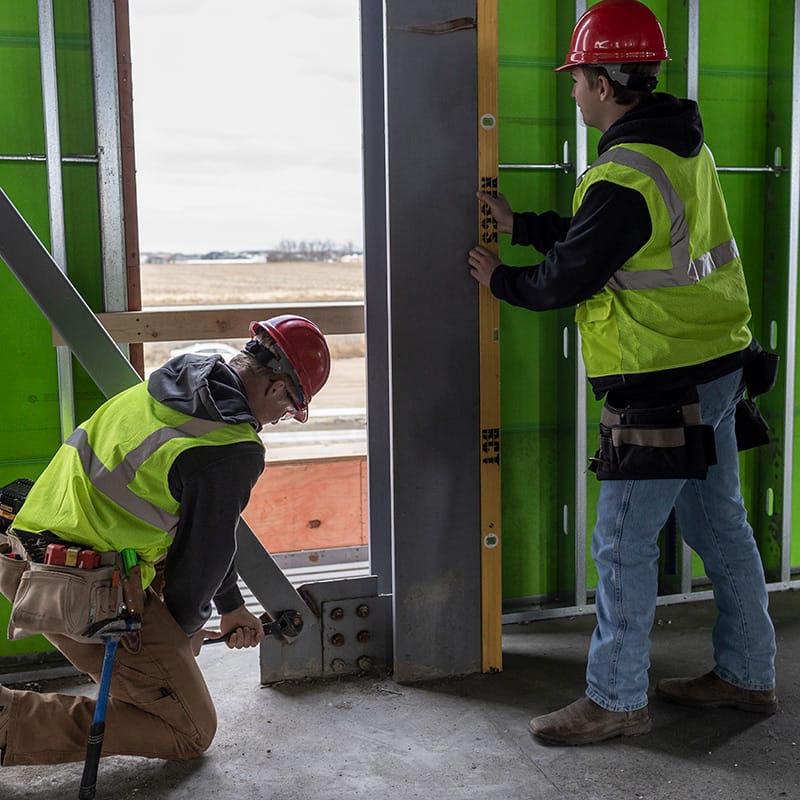 Two construction workers in red hard hats and safety vests inspecting a steel beam—one tightening a bolt with a wrench while the other holds a level against the beam.
