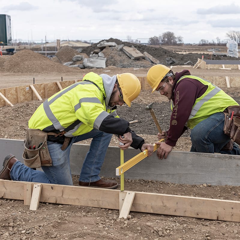 Two construction workers in yellow hard hats and safety vests measuring and securing wooden formwork at a dirt construction site.