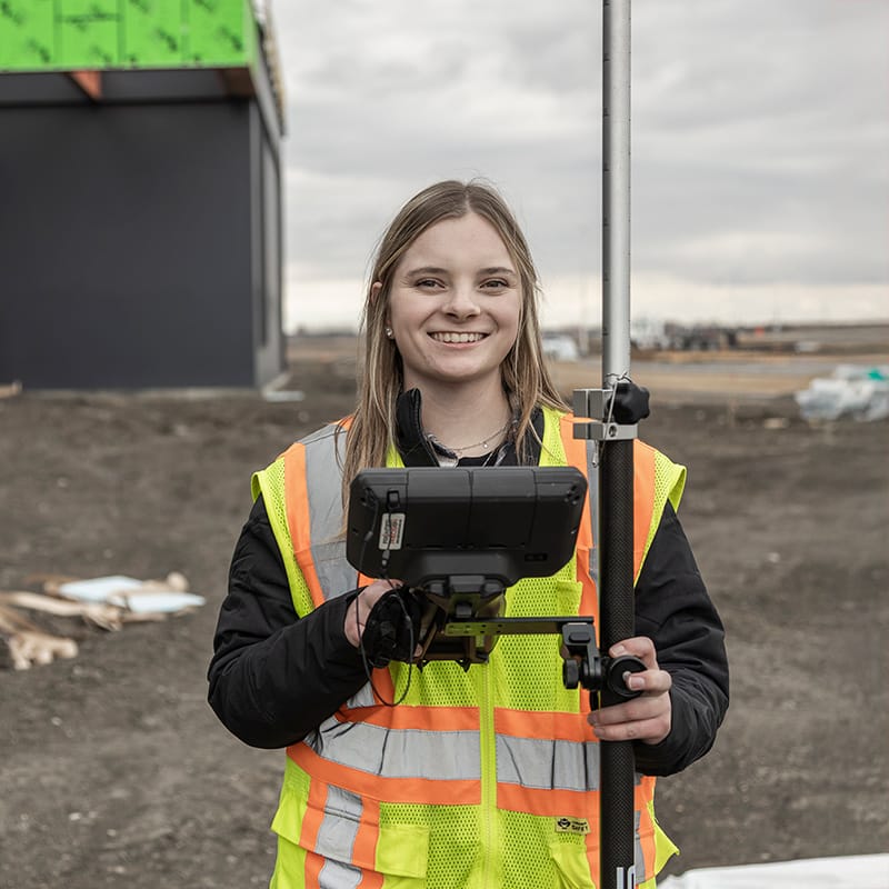 Student wearing a high-visibility safety vest smiles while holding a GPS surveying pole and monitor at an outdoor construction site.