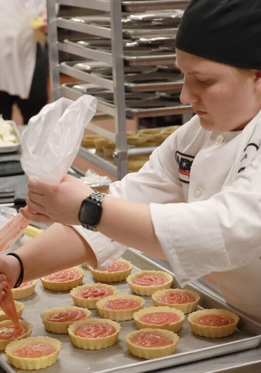 Culinary student in a white uniform and black hat piping filling into tart shells on a baking tray in a commercial kitchen.