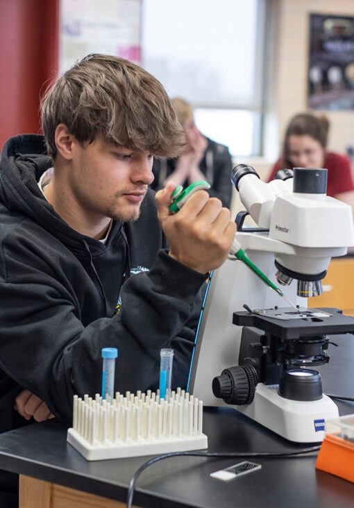 Student in a black hoodie using a pipette and microscope during a biology lab activity, with test tubes and lab equipment on the table.