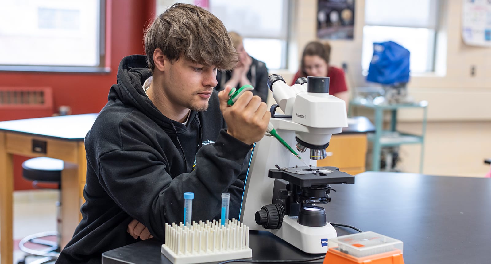 Student using a pipette and microscope in a science lab classroom with test tubes on the table