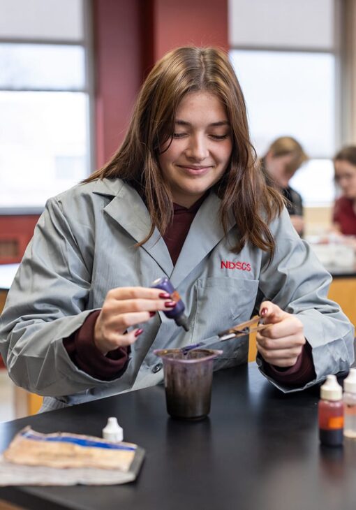 Student in an NDSCS lab coat using a dropper to add liquid to a beaker during a biotechnology experiment in a classroom.
