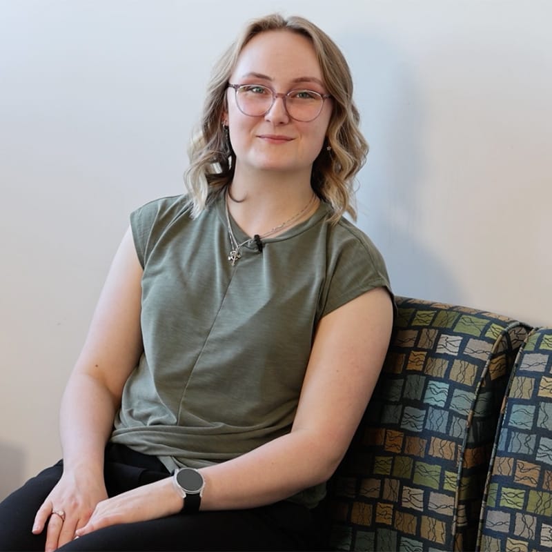 Young woman with shoulder-length blonde hair and glasses sitting in a patterned chair, smiling slightly, wearing a green top and black pants.