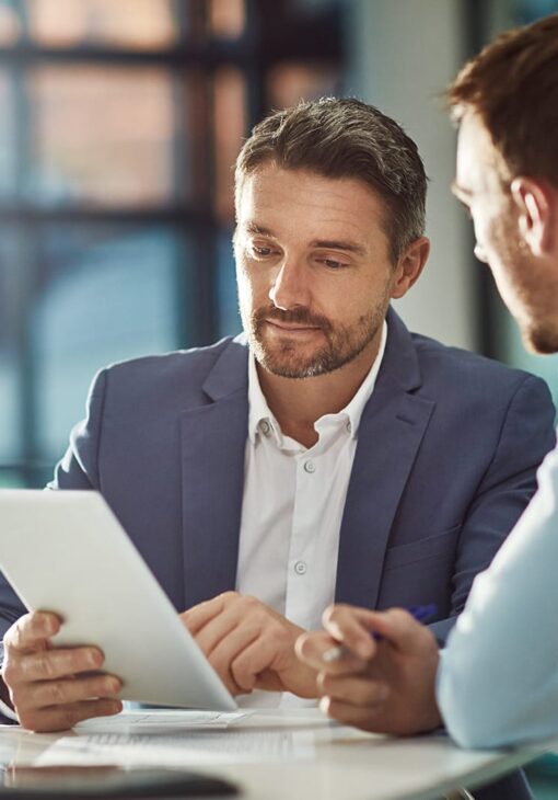Two men in business attire reviewing a document together in a modern office setting, one holding a tablet and listening intently.