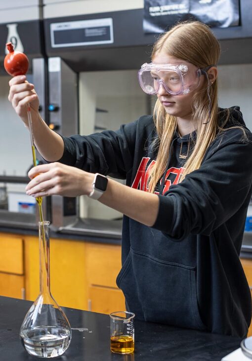 Student in safety goggles carefully transferring liquid with a pipette into a flask during a chemistry lab experiment.