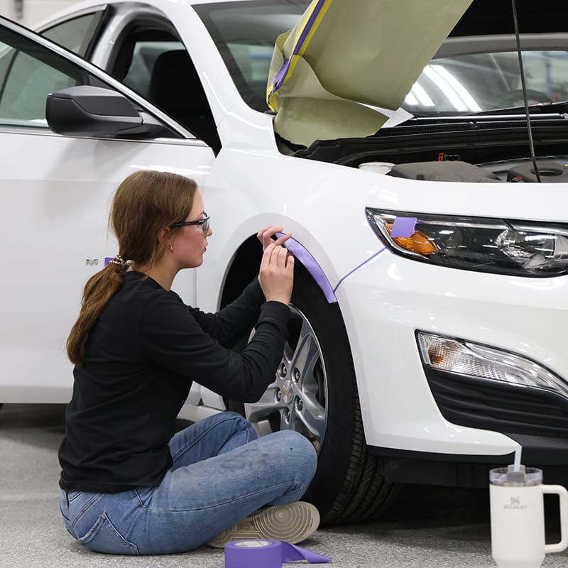Student sitting cross-legged on the floor applying purple masking tape to the fender of a white car with the hood open.