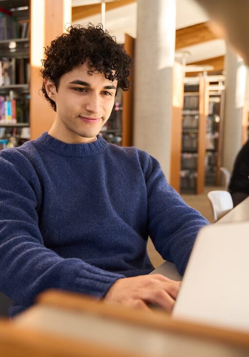 Young man in a blue sweater typing on a laptop at a library table, surrounded by bookshelves and warm natural light.