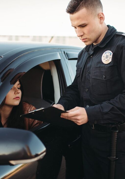 Police officer standing beside a car holding a clipboard, speaking with a woman seated in the driver’s seat during a traffic stop.