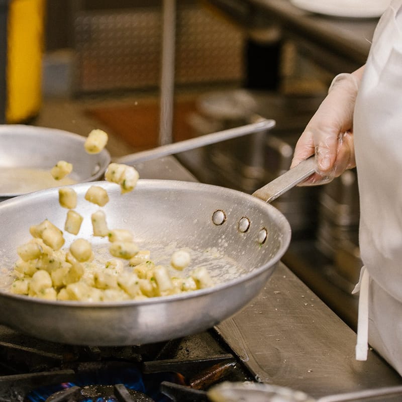 Close-up of a chef sautéing gnocchi in a large silver pan over a gas flame in a commercial kitchen.