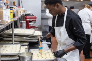 Culinary student in black chef coat and white apron brushing biscuit dough with egg wash in a commercial kitchen.