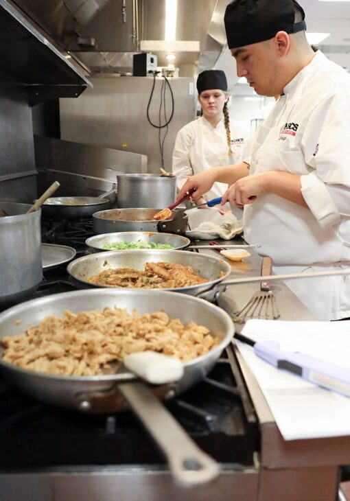 Culinary student in a white chef coat and black hat cooking multiple dishes in a commercial kitchen, with sauté pans and pots on the stovetop.