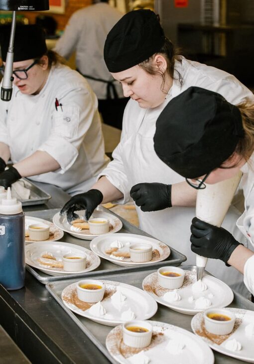 Three culinary students in white uniforms and black hats preparing dessert plates with crème brûlée, cookies, and piped whipped cream in a commercial kitchen.