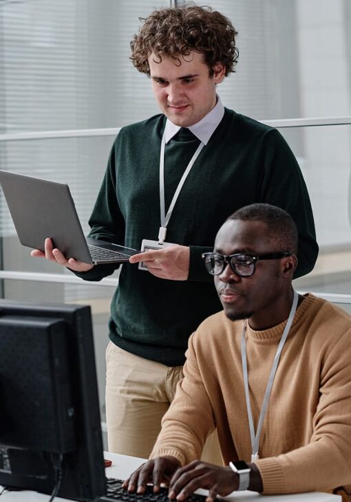 Two men working together in an office setting; one is seated typing on a desktop computer while the other stands beside him holding a laptop.