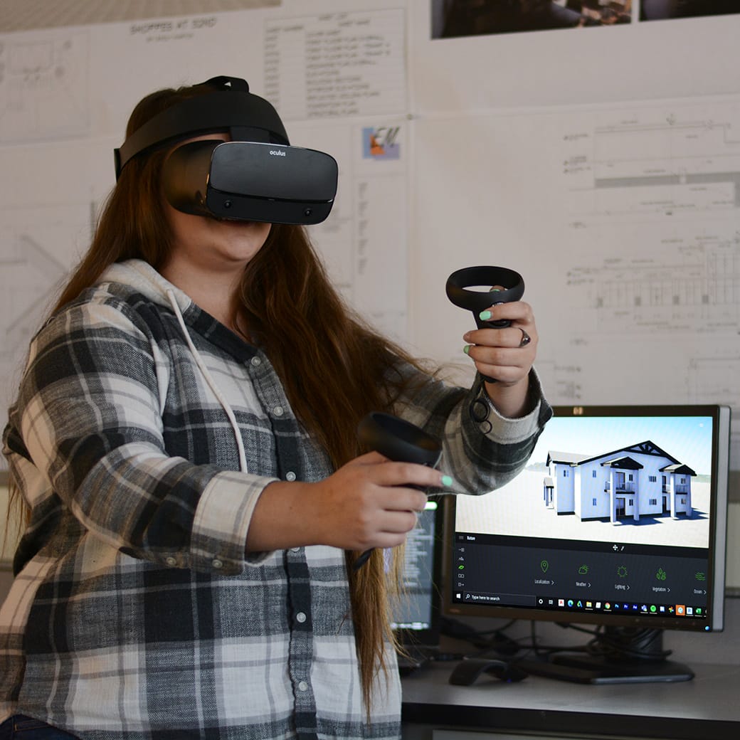 Student wearing a virtual reality headset and holding controllers, interacting with a 3D architectural model displayed on a nearby monitor.