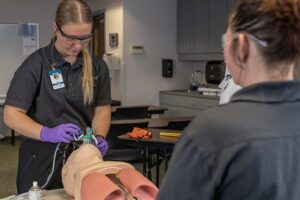 Student in safety goggles and gloves practices airway management on a medical training mannequin while another student observes.