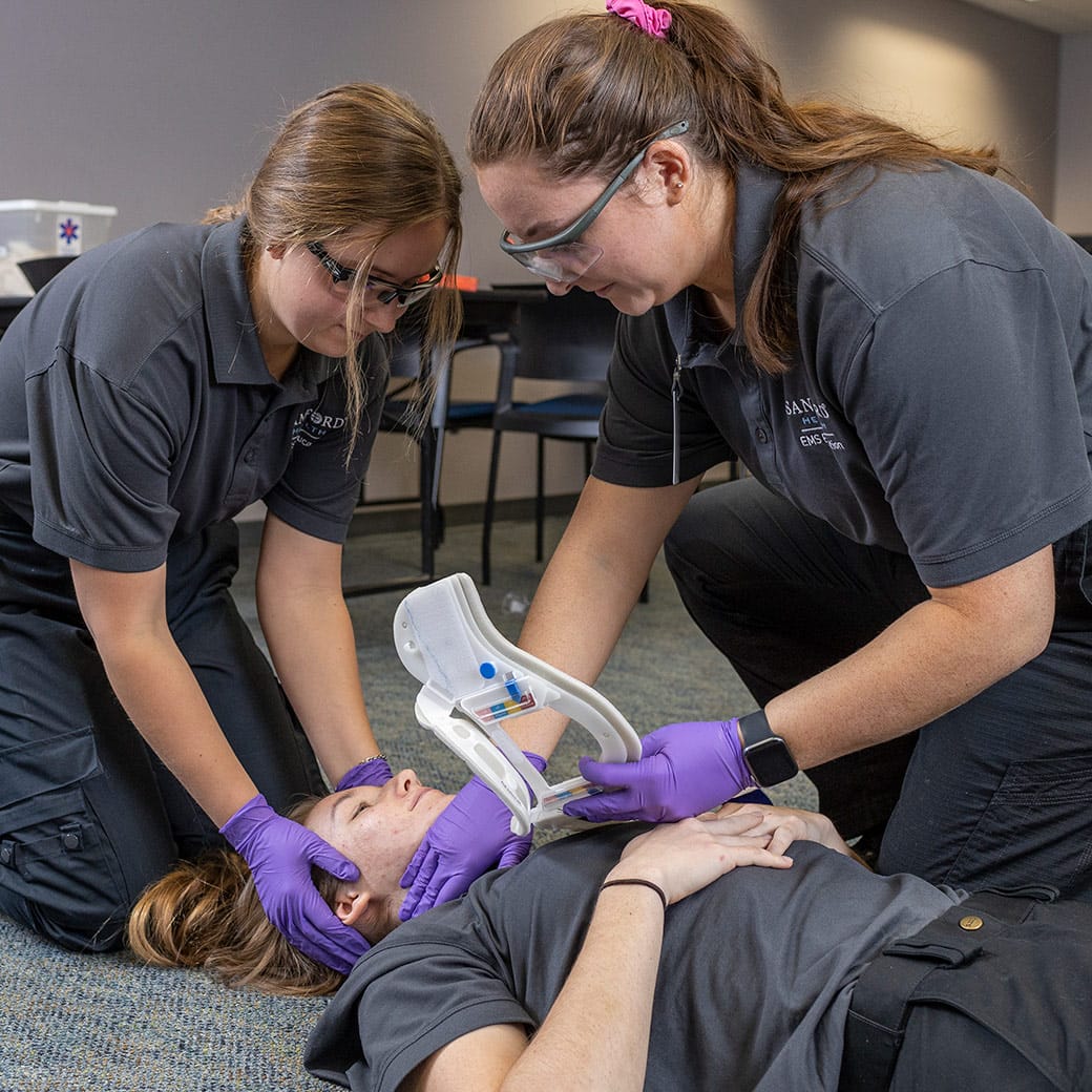 Two EMT students in uniform and safety glasses practice applying a cervical collar to a classmate lying on the floor during a medical training exercise.