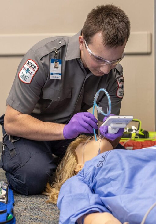EMT student wearing gloves and safety glasses practices intubation on a medical training mannequin in a classroom setting.