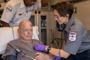 Paramedic student in gray uniform checks vital signs of an injured elderly man in an ambulance, assisted by a second paramedic in the background.
