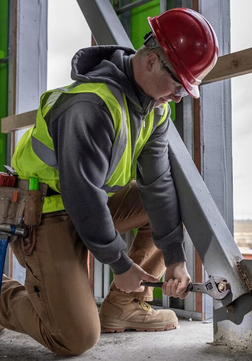 Person in a red hard hat and safety vest using a wrench to tighten a bolt on a steel beam at a construction site.