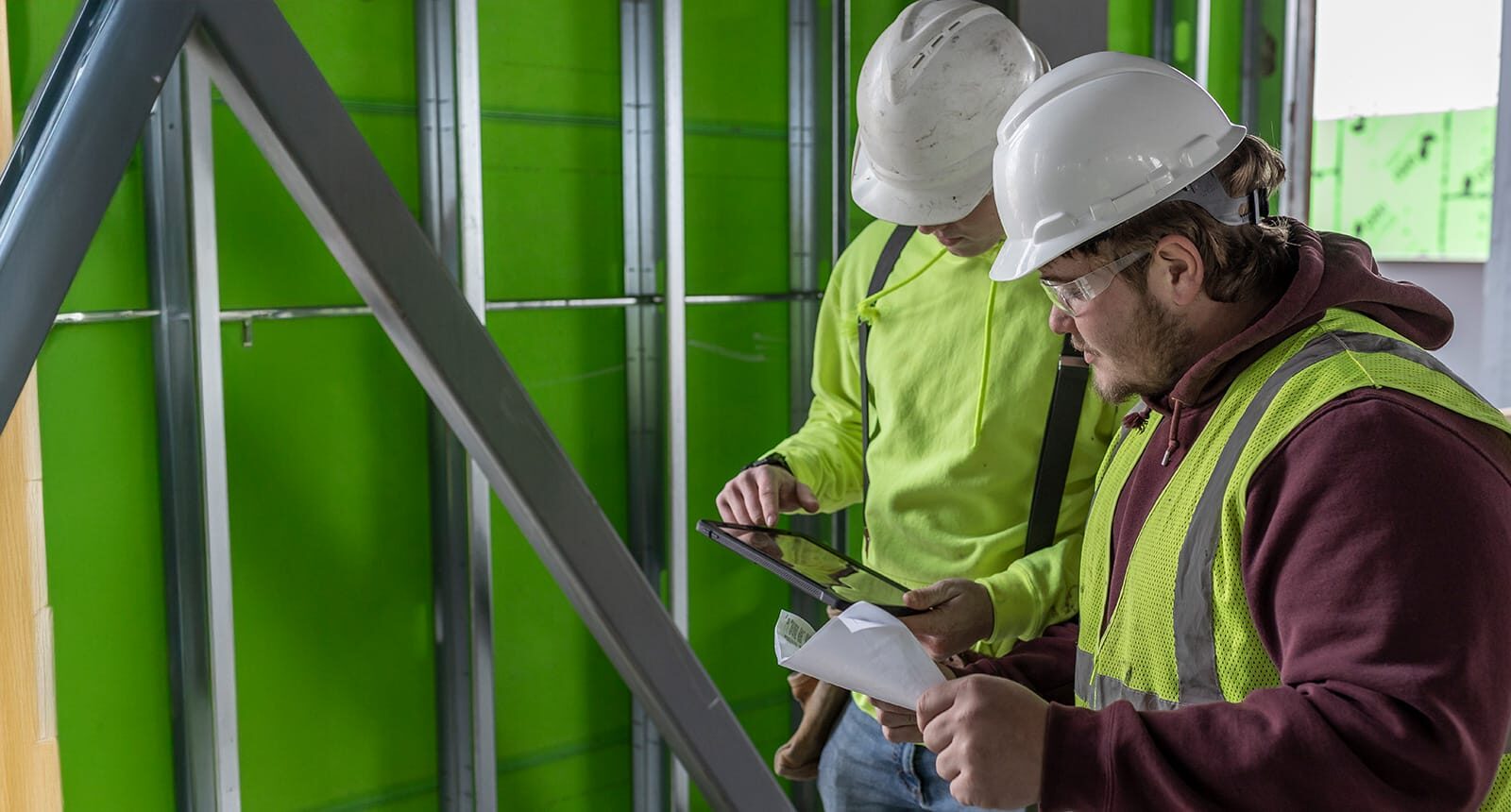 Two construction workers wearing hard hats and safety vests review documents and a tablet inside a building under construction, surrounded by metal framing and green insulation panels.