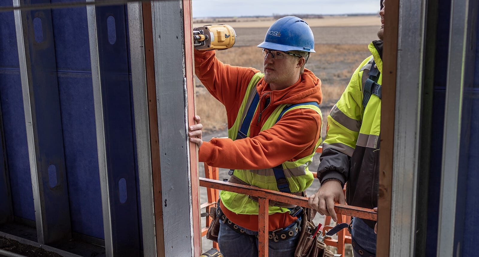 Worker in orange hoodie and safety gear using a power drill on a steel frame while standing on a construction lift, with another worker nearby.