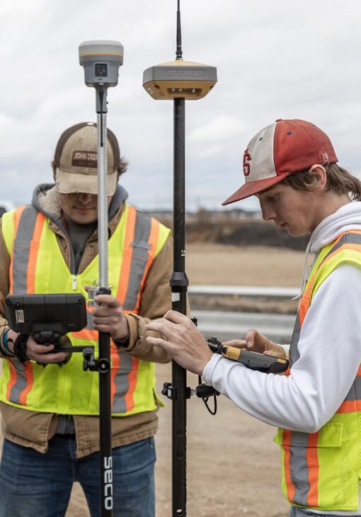 Two students in safety vests operate GPS surveying equipment on a construction site, each focused on their own device.