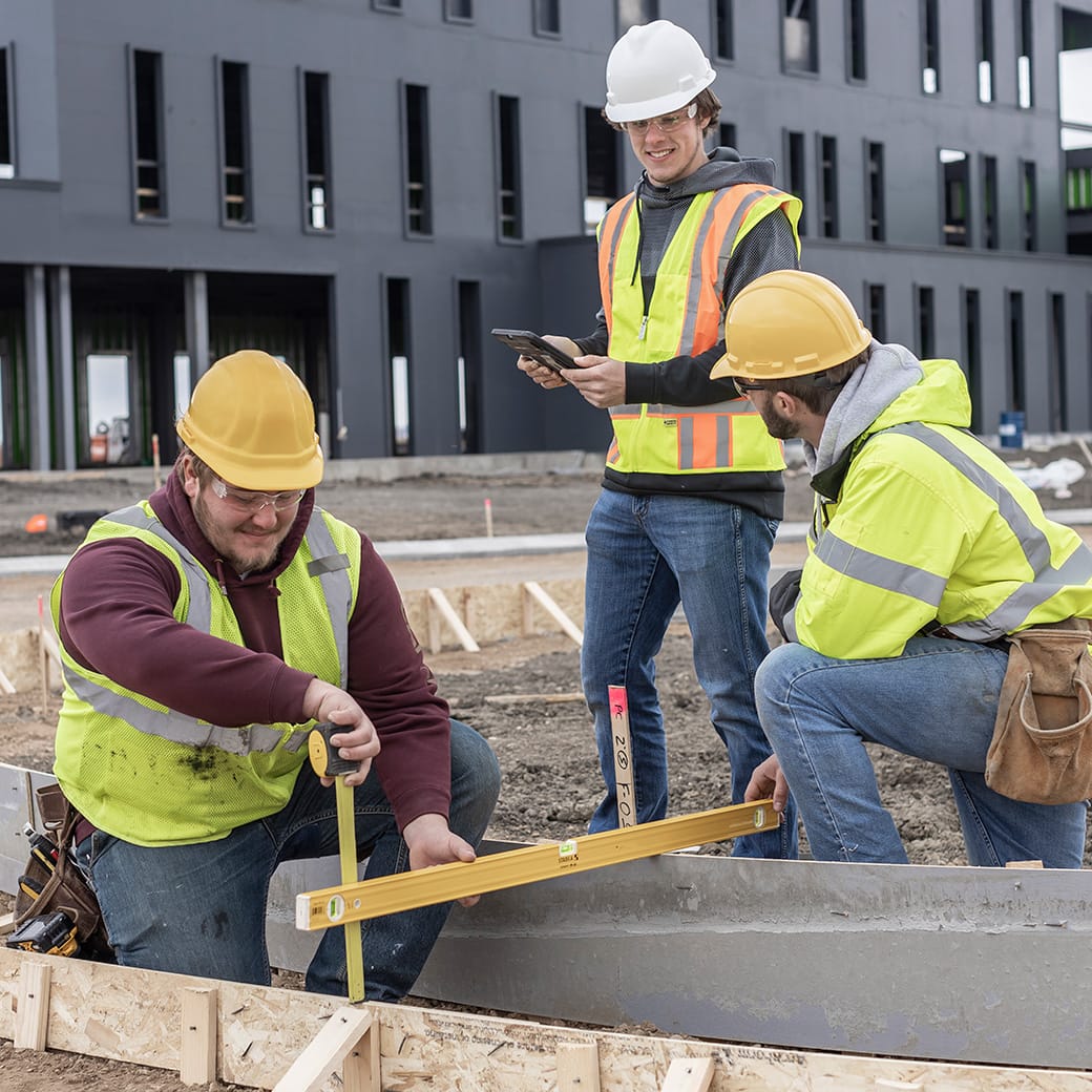 Three construction workers wearing safety gear work on-site near a modern black building; one uses a measuring tape and level while the others consult a tablet and observe the measurements.