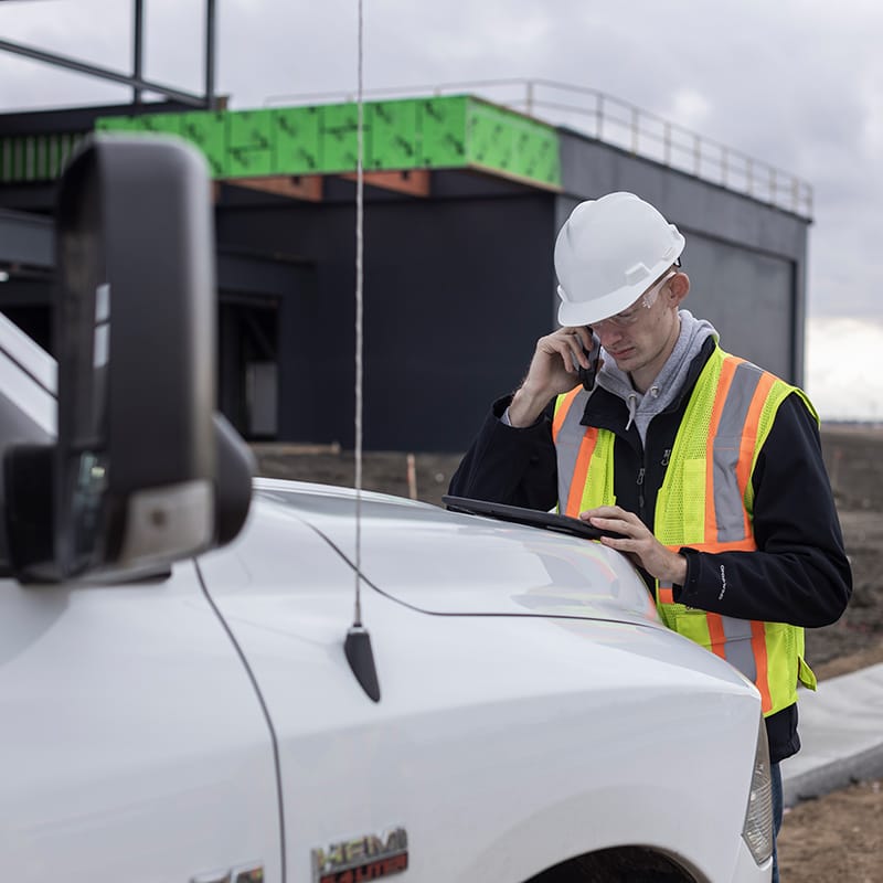 Person in a hard hat and safety vest using a phone next to a white truck at a construction site.