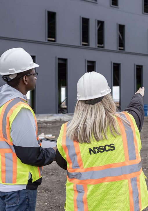 Two construction workers in safety vests and hard hats stand outside a dark gray building under construction; the woman gestures toward the structure while the man observes attentively.