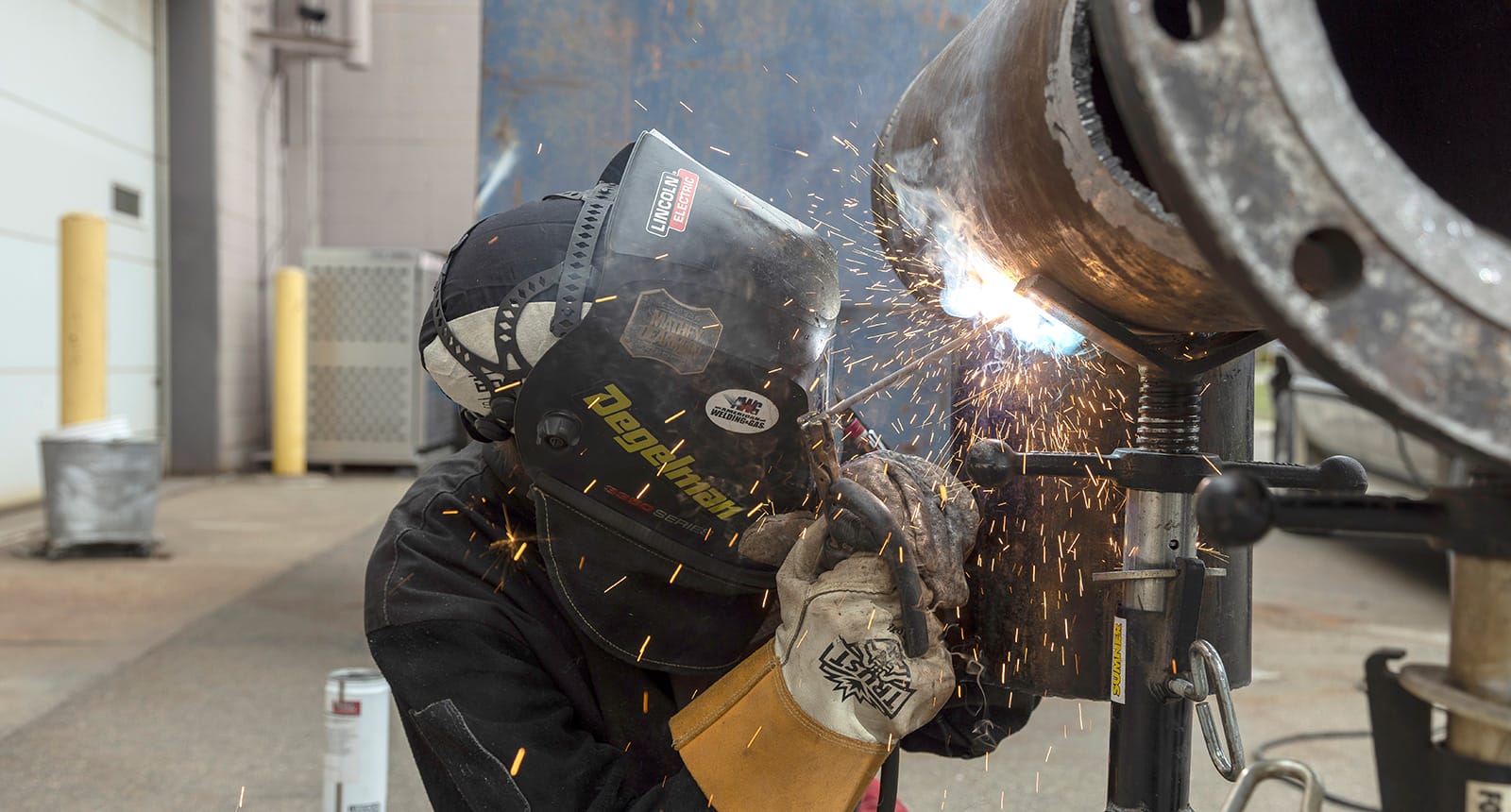 Welding student in protective gear working on a large metal pipe with sparks flying