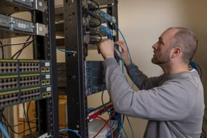 Man connecting blue network cables to a server rack in a data center or IT room.