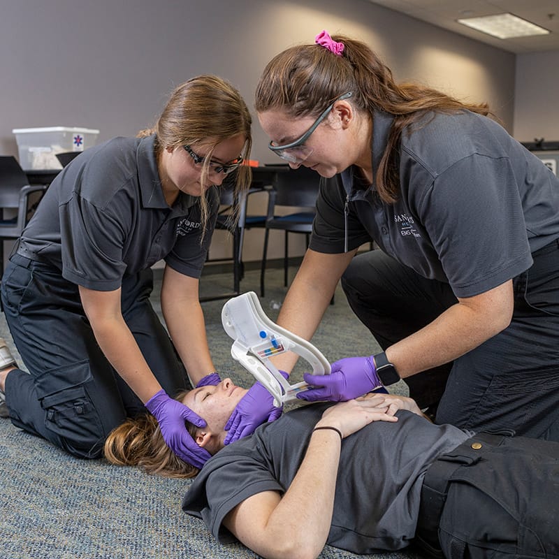 Two paramedic students in safety goggles and gloves practice airway management on a classmate lying on the floor during a training exercise.