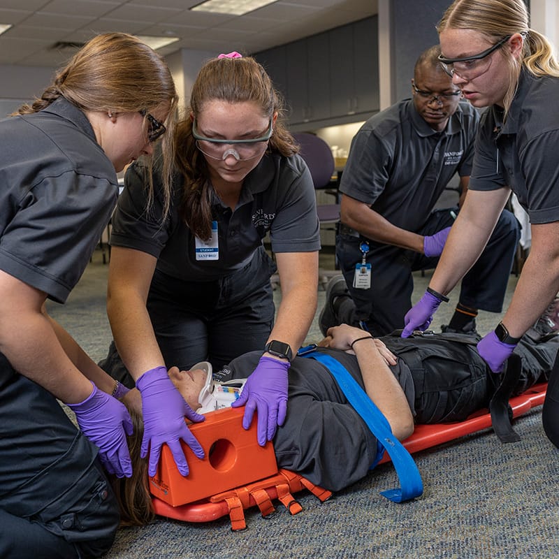 Group of EMS students in safety goggles and gloves securing a classmate to a spine board with head immobilizer during a training exercise.