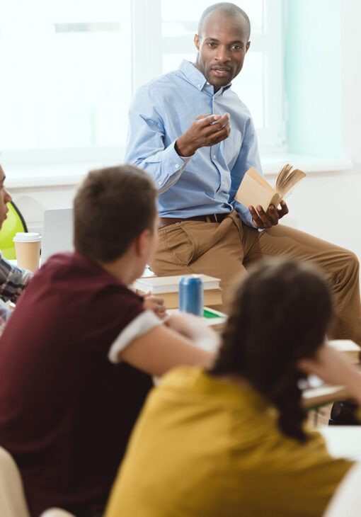 Male teacher holding an open book and gesturing while speaking to a group of college-aged students seated in a classroom.