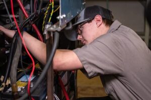 Student in safety glasses and work shirt inspecting electrical components with wires inside a large industrial machine.