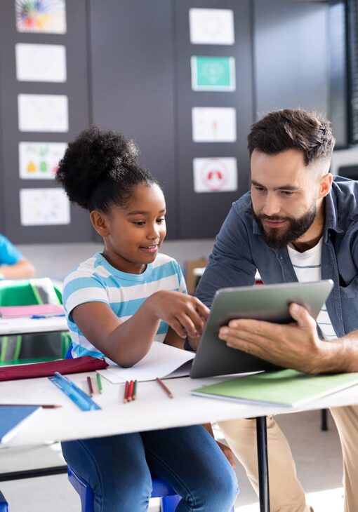 Male teacher sitting next to a young student as they look at a tablet together in a classroom filled with colorful drawings.