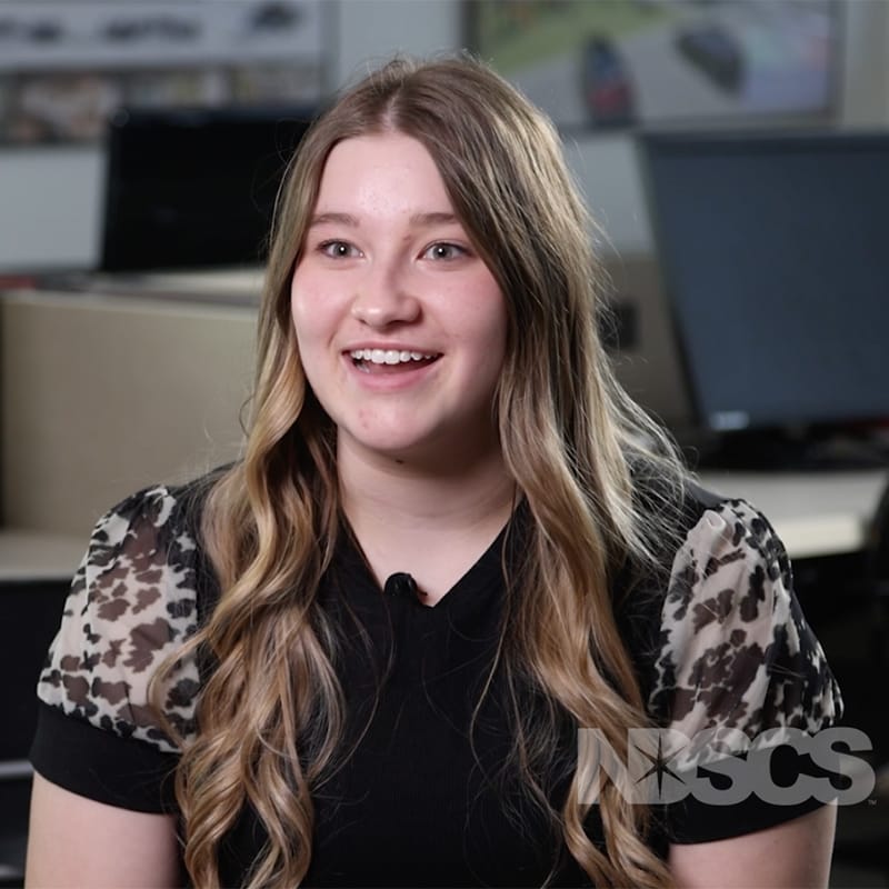 Smiling female student with long wavy hair wearing a black shirt with sheer leopard-print sleeves, seated in a computer lab with NDSCS logo in the corner.