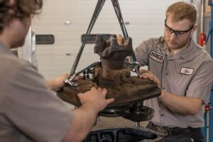 Two students in safety glasses guiding a heavy engine component into place using a shop hoist in an automotive lab.