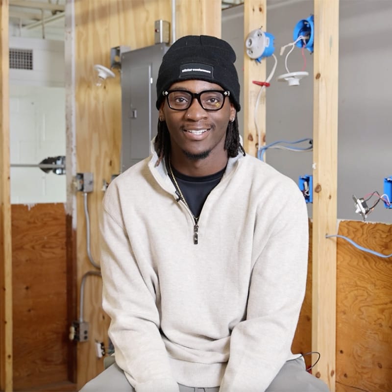 Smiling student wearing a black beanie and glasses seated in front of an electrical training setup with exposed wiring and outlet boxes.